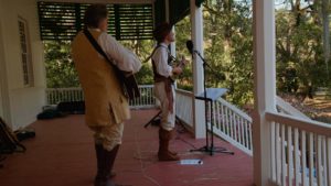 two-men-wearing-historical-costumes-playing-music-standing-on-red-porch