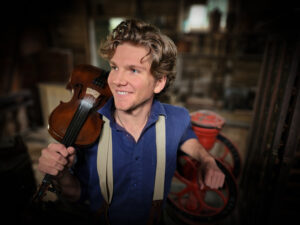 blonde haired smiling man wearing blue shirt and white suspenders holding violin on shoulder