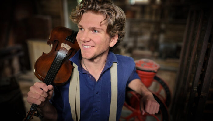 blonde haired smiling man wearing blue shirt and white suspenders holding violin on shoulder