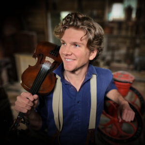 blonde haired smiling man wearing blue shirt and white suspenders holding violin on shoulder